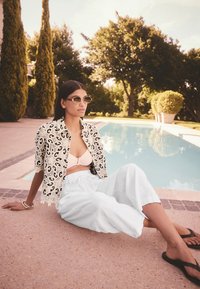 Giacca corta a motivo bianco e nero sopra una maglietta rosa chiaro e pantaloni bianchi larghi; occhiali da sole rotondi; seduta accanto a una piscina.