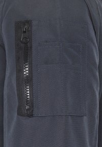 Tissu bleu marine à finition texturée, avec une poche zippée noire proéminente et une petite poche plate rectangulaire au-dessus.