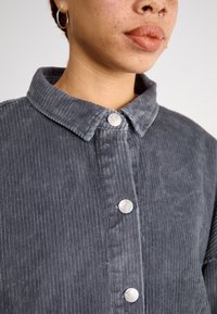 Hnedá kordurová košeľa s golierom, s predným zapínaním na strieborné gombíky a textúrovaným prúžkovaným vzorom.