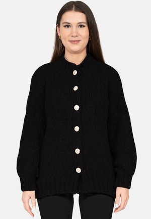 Vrouw met lang bruin haar die een zwarte gebreide cardigan met zes grote gouden knopen draagt, staand tegen een eenvoudige lichte achtergrond.