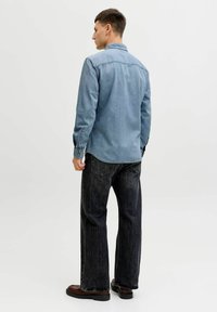 Jeanshemd in Hellblau, mit spitzem Kragen, geknöpften Manschetten und abgerundetem Saum. Kombiniert mit ausgewaschenen schwarzen Jeans und dunklen Schuhen.