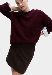 Bordeaux wollen trui met een relaxte pasvorm, ronde halslijn en verlaagde schouders, gelaagd over een witte blouse met kraag en een bruine rok.