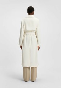 Langer, hellcremefarbener Trenchcoat mit tailliertem Schnitt und Gürtel in der Taille sowie einem leicht ausgestellten Rock. Verfügt über einen Reverskragen und lange Ärmel.