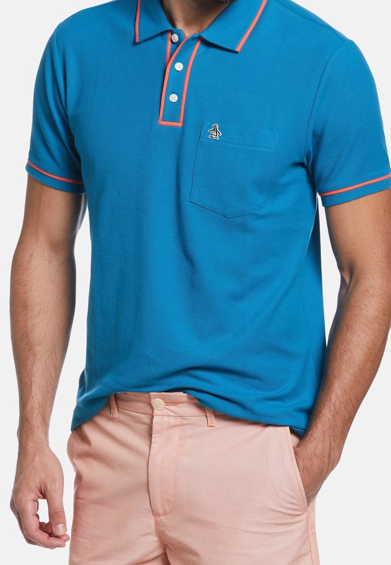 Homme portant un polo bleu avec bordure rouge et logo sur la poche, associé à un pantalon rose clair, une main dans la poche, sur fond blanc.