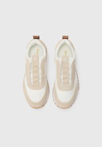 Un paio di sneaker slip-on beige e bianche con dettagli in suede e linguette, viste dall'alto su uno sfondo bianco.