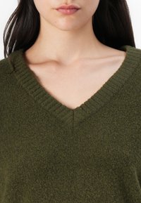 Olivenfarget strikkegenser med teksturert finish, med dyp V-hals og ribbestrikkede mansjetter. Stoffet ser mykt og varmt ut.