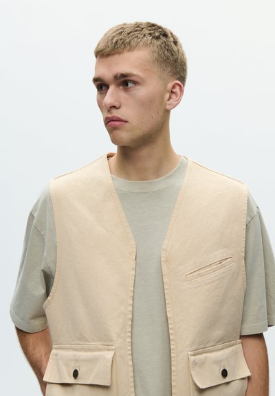 Młody mężczyzna z krótkimi blond włosami, ubrany w beżową kamizelkę użytkową na jasnoszarej koszulce, patrzący w lewo na jednolitym tle.