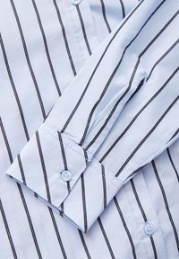 Manche de chemise bleu clair avec des rayures verticales noires et un poignet boutonné, posée sur un corps de chemise rayé assorti.