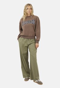 Donna che indossa un maglione di pile marrone con la scritta "COLORADO", pantaloni larghi oliva e sneakers marroni, in piedi davanti a uno sfondo neutro.