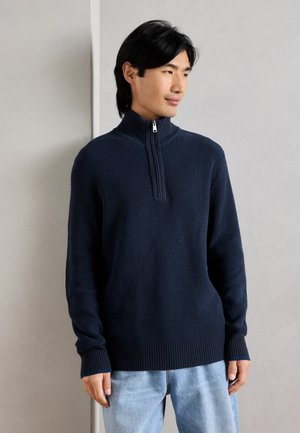 Marineblauer Rippstrickpullover mit hohem Kragen und Reißverschluss. Lange Ärmel und lockere Passform, aus strukturiertem Stoff mit gerippten Bündchen.