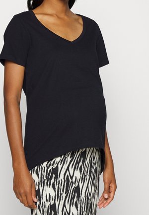 Zwangere persoon draagt een zwart V-hals top met korte mouwen en een zwart-witte rok met patroon, afbeelding is bij de schouders en dijen bijgesneden.