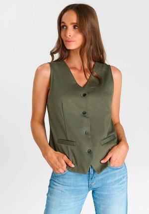 Vrouw met lang bruin haar, draagt een olijfgroene mouwloze vest met knopen en lichtblauwe jeans, handen in de zakken, kijkt naar links.