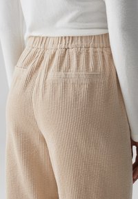 Beige corduroy broek met verticale ribbels, elastische tailleband en twee achterzakken, gezien vanaf de zijkant en iets van achteren.