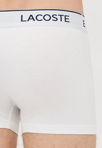 Boxer briefs bianchi aderenti con logo "Lacoste" nero sulla vita elastica indossati da un uomo in piedi.