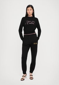 Svart strikkegenser med rosa brodert "Versace Jeans Couture"-tekst, kombinert med svarte joggebukser med brodert logo. Hæler.