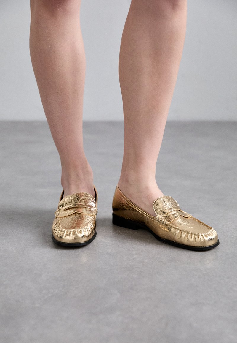 Mocassins en cuir doré avec un bout arrondi, des coutures décoratives et une finition texturée. Semelle en caoutchouc noir et design minimaliste.