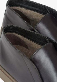 Ciemnobrązowe skórzane buty z gładką fakturą, zaokrąglonym kształtem i miękkim filcowym wykończeniem. Charakteryzują się minimalnym przeszyciem i niskoprofilową podeszwą.