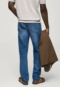 Vaqueros de mezclilla azul con corte recto, que presentan bolsillos traseros y costuras visibles, combinados con una chaqueta exterior marrón sostenida en una mano.