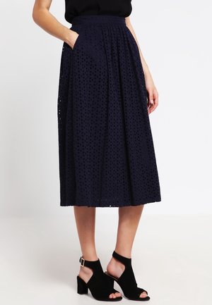 Kobieta w granatowej spódnicy midi z kwiatowym wzorem dżetów, czarnym topie bez rękawów oraz czarnych sandałach na obcasie z odkrytymi palcami, stojąca z jedną ręką w kieszeni.