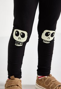 Sorte leggins med to hvide skull-graphics på knæene, lavet i et strækbart stof, parret med brune velcro sneakers.