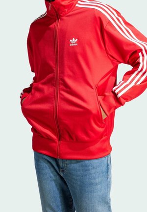 Personne portant une veste de sport rouge Adidas avec des bandes blanches sur les manches et un logo sur la poitrine, associée à un jean bleu, mains dans les poches.