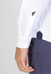 Chemise blanche à manches longues avec un petit cheval navy brodé sur le poignet, assortie à un pantalon bleu foncé. Tissu lisse et coupe ajustée.