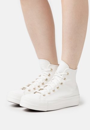 Gambe che indossano scarpe da ginnastica alte in tela bianca pulita con lacci bianchi e suole di gomma spesse su uno sfondo semplice.