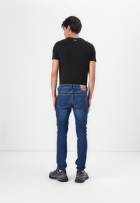 Camiseta negra de cuello redondo y jeans de denim azul. Los jeans cuentan con dos bolsillos traseros y una etiqueta de cuero marrón. El modelo lleva zapatillas gris.