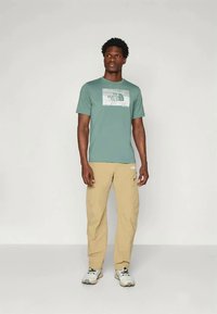 Maglietta verde chiaro con design grafico e logo sul petto, abbinata a pantaloni beige chiaro e scarpe sportive grigie.
