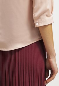 Jasnoróżowa, przezroczysta bluzka z podwiniętymi rękawami i zapięciem na guziki, zestawiona z plisowaną bordową spódnicą, podkreślająca delikatne tekstury i uzupełniające kolory.