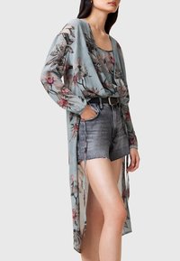 Kimono floreale in tessuto azzurro tenue, con maniche lunghe, design fluido e dettaglio a spacco. Indossato sopra pantaloncini di denim con bordi sfrangiati.