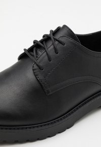 Black leather shoe with a smooth texture, rounded toe, and lace-up design. Features subtle stitching and a sturdy rubber sole with treads.
