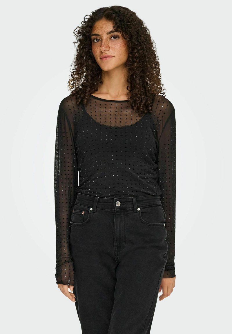Sort langærmet top med prikket mesh-stof, der har en rund halsudskæring og tætsiddende design, parret med høj talje sorte jeans.