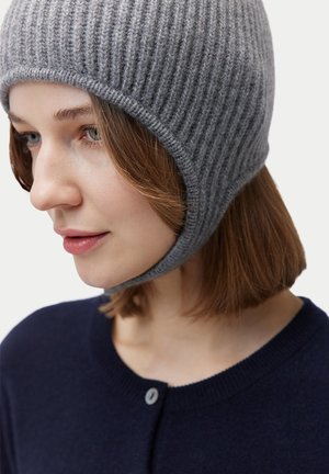 Junge Frau mit schulterlangen braunen Haaren, die eine gerippte graue Strickmütze und einen dunkelblauen, geknöpften Pullover trägt.