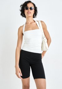 Bela zgornja majica na trakove s kvadratnim izrezom, teksturirana tkanina, v kombinaciji s črnimi kratkimi kolesarskimi hlačami. Dodatki vključujejo majhna črna sončna očala in kremasto torbico.