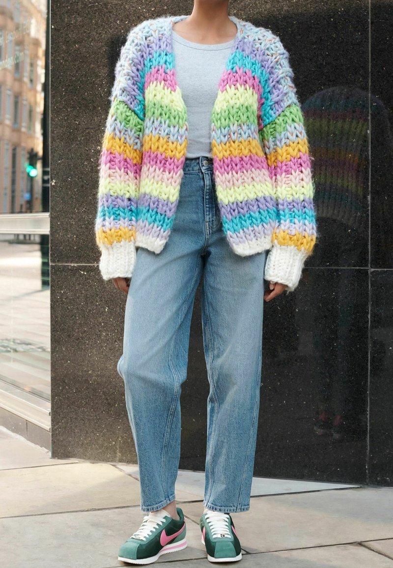 Personne portant un cardigan en grosse maille arc-en-ciel pastel, un haut gris clair, un jean bleu taille haute et des baskets vertes et roses, debout en plein air.