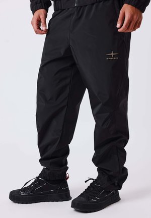 Man wearing black pants with elastic cuffs and black sneakers with thick soles, standing against a plain light background.