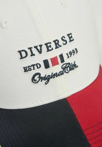 Czapka baseballowa z białym przodem, czerwonymi i granatowymi panelami. Ozdobiona haftowanym napisem "DIVERSE ESTD 1993 OriginalClth" oraz małą grafiką flagi.
