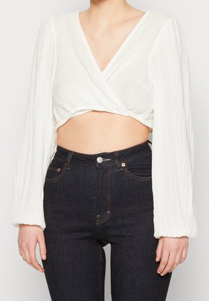 Kobieta ubrana w biały, długi rękaw, krótki top z wiązaniem, zestawiony z ciemnoniebieskimi, wysokim stanem dżinsami, stojąca na jednolitym tle.