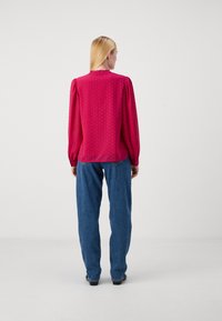 Blusa fucsia con maniche lunghe a sbuffo, scollo rotondo e pois in rilievo. Abbinata a jeans blu larghi, vista da dietro.