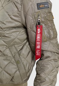 Olivgrüne gesteppte Bomberjacke mit Reißverschlusstasche, gerippten Bündchen und rotem "Remove Before Flight"-Anhänger am Reißverschluss.