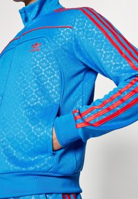 Modrá bunda od teplákové soupravy s červenými pruhy, vyrobená z hladkého materiálu; má zipový límec, logo Adidas a vzor s trojlístkovými logy.
