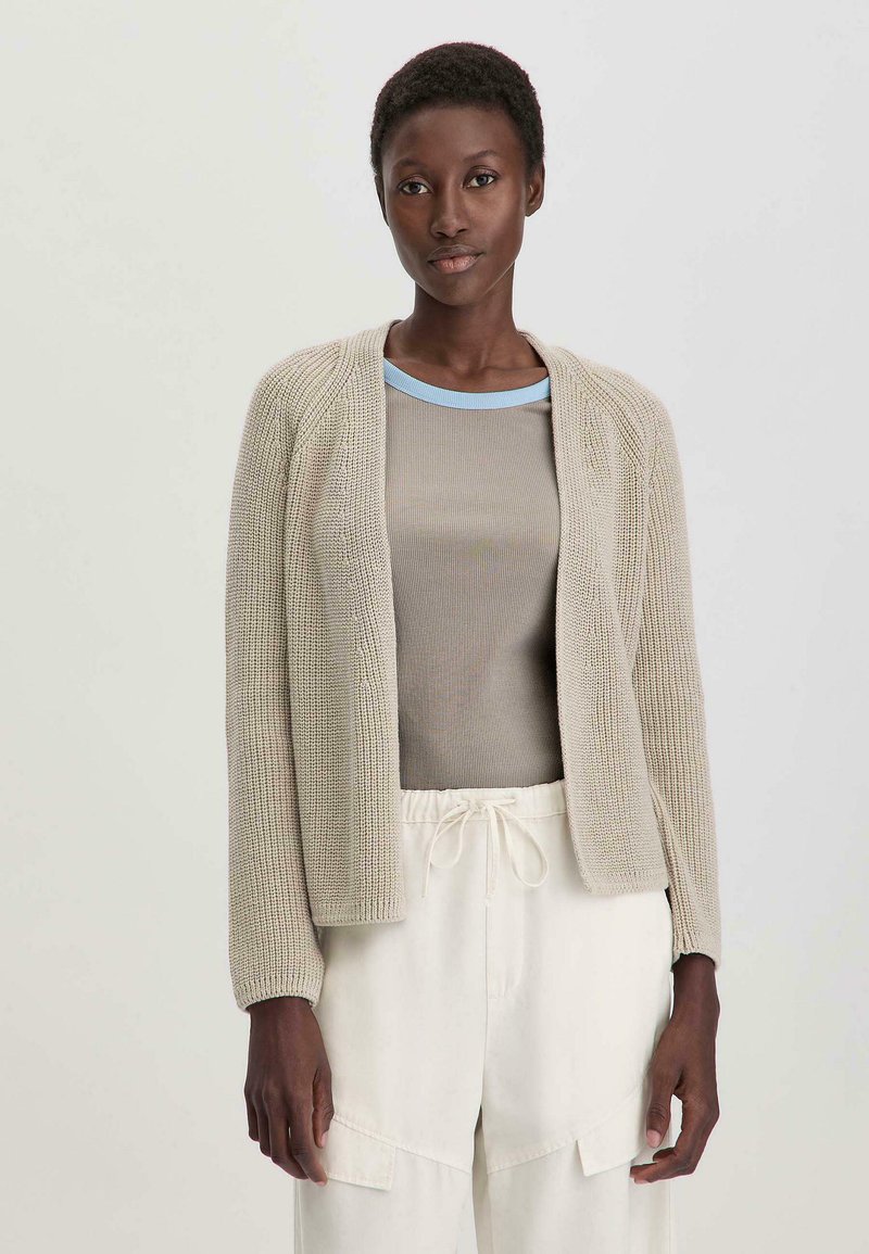 Cardigan de punto de color beige claro con abertura frontal, mangas largas; combinado con una blusa gris claro con cuellos azules y pantalones blancos con cordón.