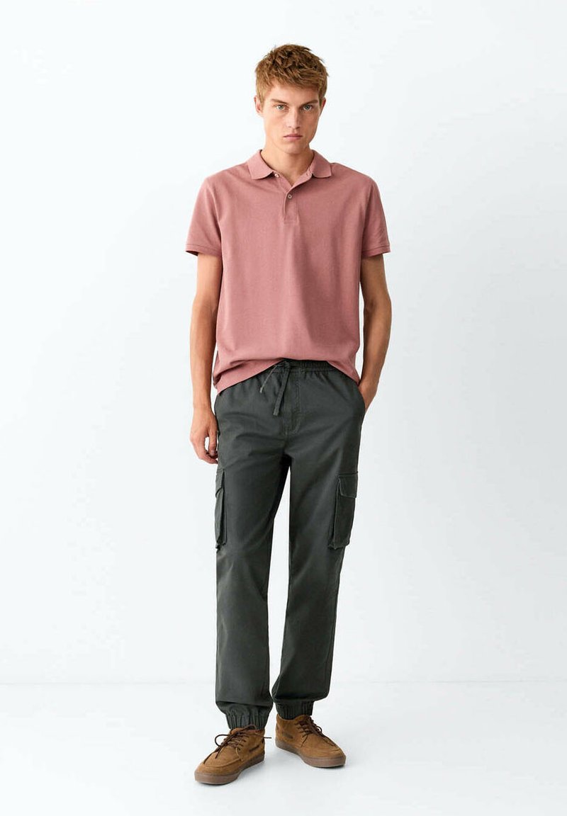 Jeune homme debout les mains dans les poches, portant un polo rose poussiéreux, un pantalon cargo foncé et des chaussures décontractées marron sur fond blanc.
