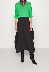 Top in maglia verde con colletto, abbinato a una lunga gonna nera con cuciture curve. La modella indossa stivali neri con tacco e una sciarpa a fantasia.