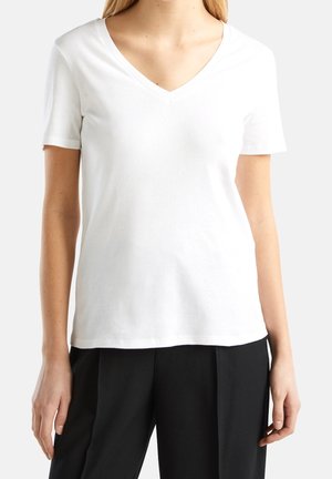 Kvinde iført almindelig hvid V-hals kortærmet T-shirt og sorte plisserede bukser, vist fra nakke til hofter på lys baggrund.