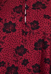 Pattern floreale scuro su uno sfondo di tessuto rosso, con bottoni neri e dettagli a pois lungo la pattina.