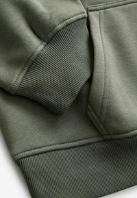 Gros plan sur le poignet et la poche d'une manche de sweat-shirt vert olive avec tissu tricoté côtelé et détails de coutures visibles.
