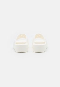 Ett par vita Crocs-sandaler sedd från baksidan, som visar ventilationshål och texturerade sulor på en enfärgad bakgrund.