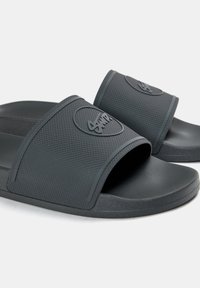 Svarta gummisandaler med strukturerade remmar. Har en logotyp framtill och en platt sula. Enkel design och lätt att ta på.